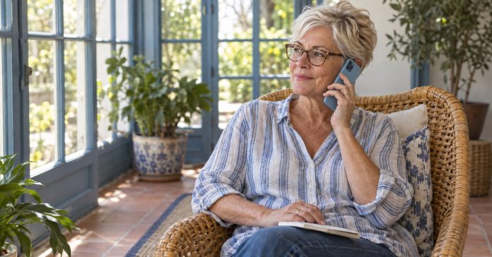 Woman in her fifties seated in a sunroom with a modern light-blue smartphone at her right ear and a notebook on her lap.